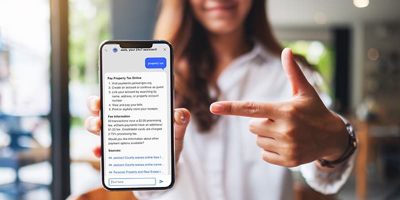 A person holds a smartphone displaying a webpage about paying property tax online, with their other hand pointing at the screen.