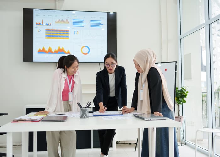 Three businesswomen collaborating on data report with dashboard displayed on screen in modern office setting