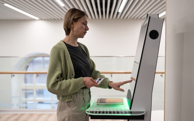A person stands in front of a touchscreen, self-service kiosk while holding their library card.