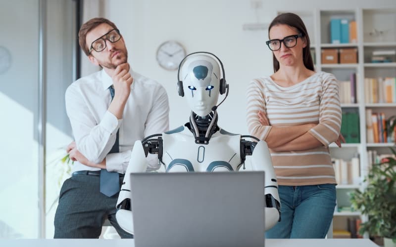 A humanoid robot sits confidently at a desk with a laptop, while two colleagues standing behind it appear uncertain.
