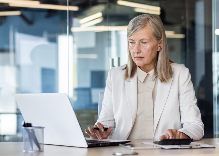 Professional woman in a white blazer working on a laptop at a modern office desk, appearing focused and engaged.