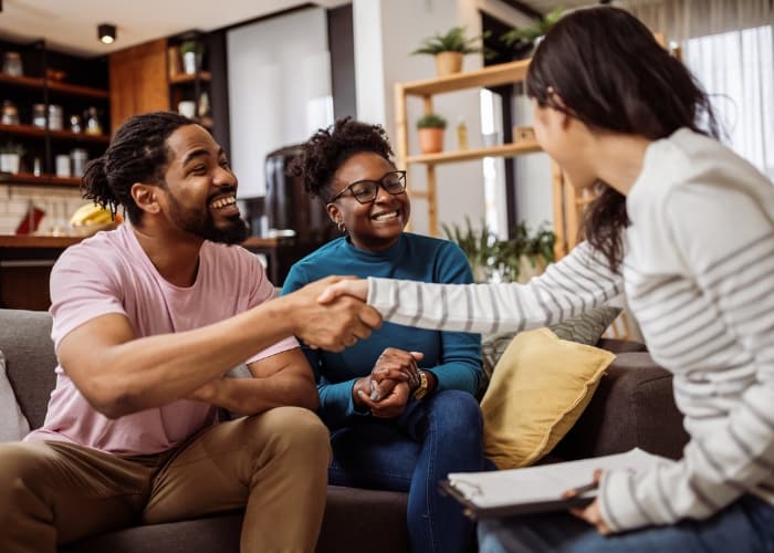 A couple of tenants sit on a couch in a living room, shaking hands with a landlord holding a clipboard.