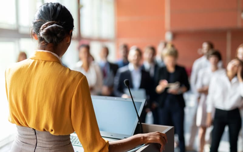 A woman in a yellow sweater stands at a podium and speaks to a group of people.