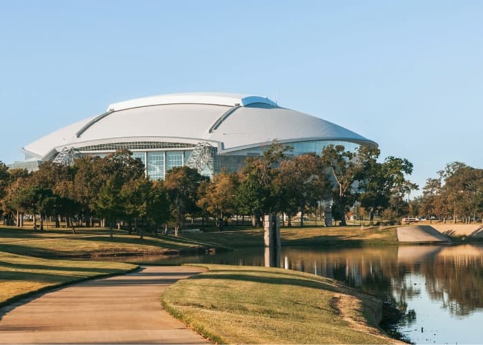 Modern domed stadium in Arlington, Texas, surrounded by trees and a reflective pond.