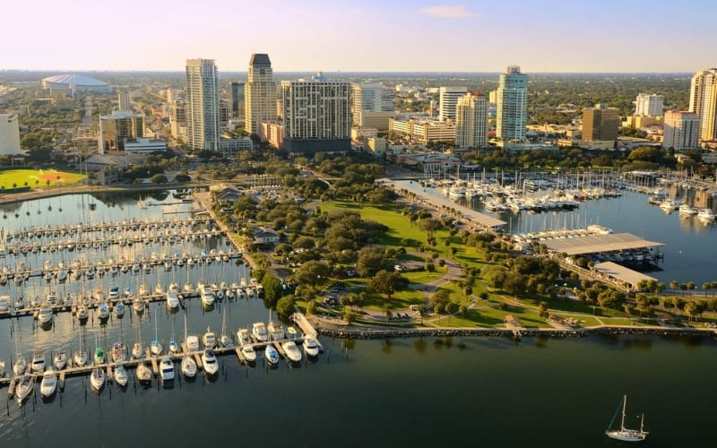Aerial view of downtown St. Petersburg, Florida, featuring a marina filled with boats, lush green parks and a skyline of modern high-rise buildings.