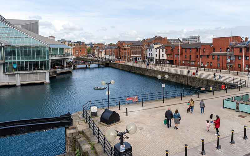 Canal scene with brick and glass buildings, people walking along the promenade, and a small boat on the water under a partly cloudy sky.