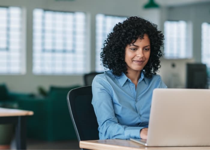 A person with curly hair, wearing a blue shirt, sits at a desk working on a laptop in a modern office with large windows in the background.