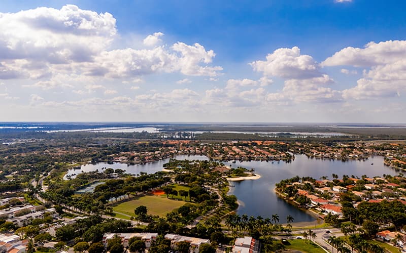 Aerial view of a suburban neighborhood in Doral, Florida, with houses surrounding a lake, green spaces, and trees under a partly cloudy sky.