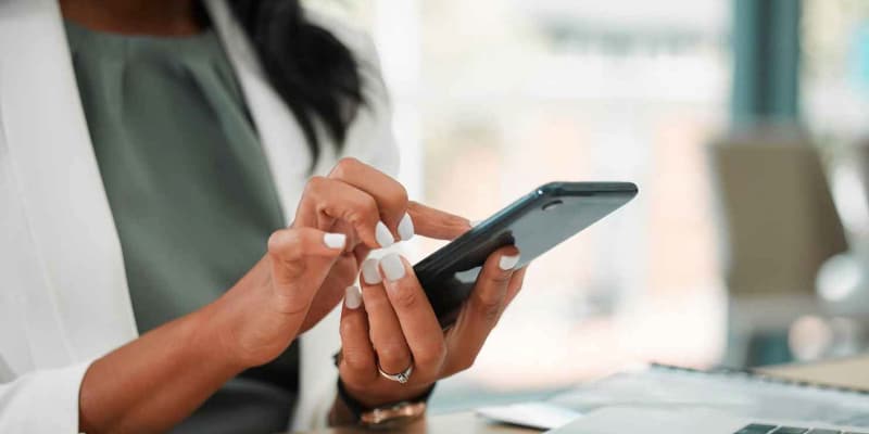 A person with long white nails using a smartphone, with a laptop and scattered papers on a desk in the background.