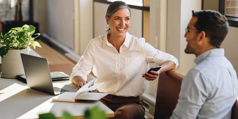Two people sitting at a table in an office, with a smartphone, open book, laptop, and potted plant on the table.