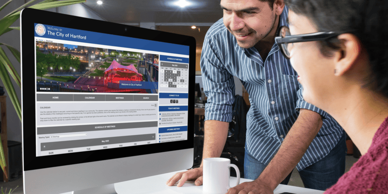 Two people look at a computer monitor open to the City of Hartford meeting schedule web page.